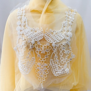 May include: A pale yellow sheer fabric garment featuring an intricate white beaded applique across the chest. The embellishment consists of small white pearls and clear crystals arranged in geometric patterns, including lattice work and dangling accents.
