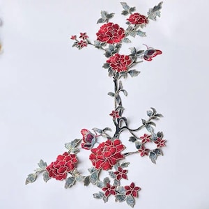 Könnte beinhalten: Ein gestickter Aufnäher mit einem Zweig, der leuchtend rote Pfingstrosen, kleinere rote Blüten und graugrüne Blätter zeigt. Zwei Schmetterlinge mit rot-blauen Flügeln sind in das florale Design integriert, das mit detailliertem Garn gearbeitet ist.