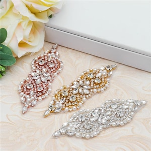 Puede incluir: Tres apliques decorativos con cristales facetados transparentes y pequeñas perlas blancas dispuestas en complejos patrones florales. El conjunto incluye una base de metal en oro rosa, oro y plata, cada una con un diseño simétrico.