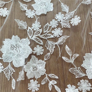 Puede incluir: Primer plano de tela de encaje floral blanco. El encaje presenta bordados detallados de varios dise&ntilde;os de flores y hojas. Los delicados patrones se destacan sobre un fondo transparente y de color claro, creando una apariencia texturizada y elegante.