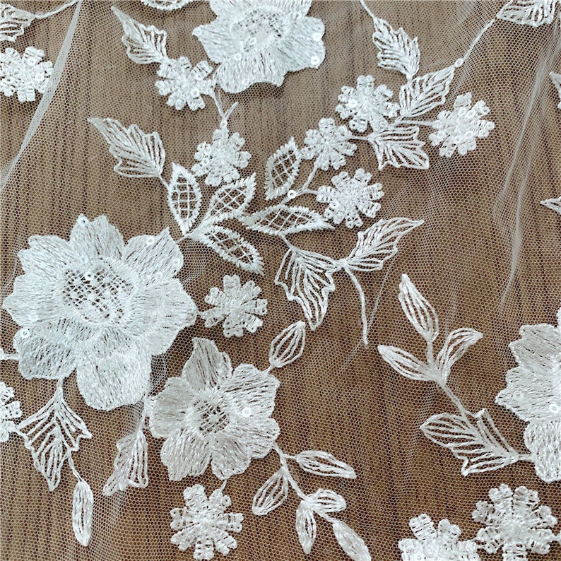 Puede incluir: Primer plano de tela de encaje floral blanco. El encaje presenta bordados detallados de varios dise&ntilde;os de flores y hojas. Los delicados patrones se destacan sobre un fondo transparente y de color claro, creando una apariencia texturizada y elegante.