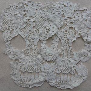 May include: Two white lace skull shapes, with intricate floral designs, are displayed on a beige background.