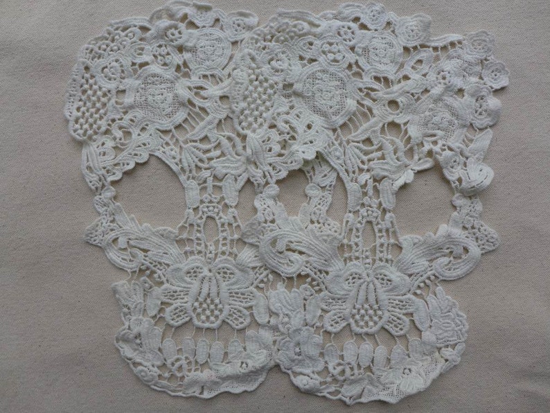May include: Two white lace skull shapes, with intricate floral designs, are displayed on a beige background.