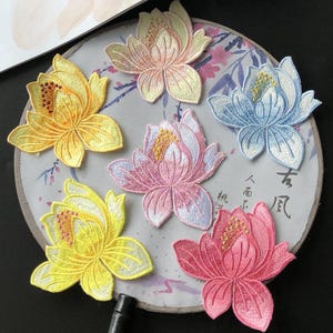 May include: Embroidered lotus flower patches in shades of yellow, pink, and blue. The patches are arranged on a decorative fan with Asian-inspired artwork. The patches have detailed stitching and a textured appearance.