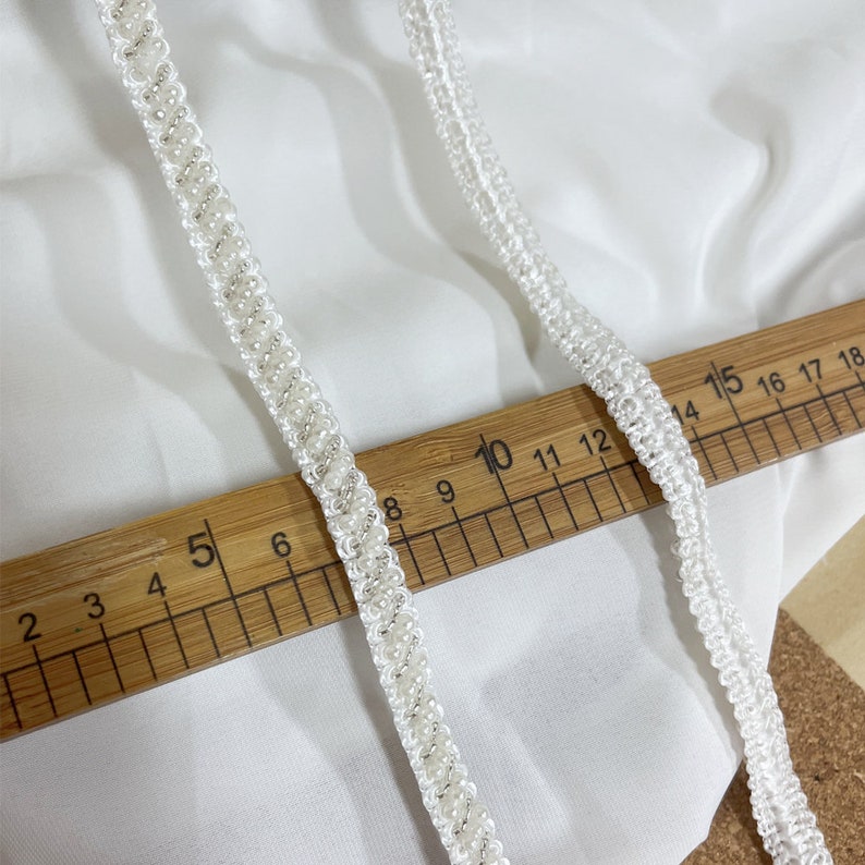 May include: Two white beaded trims with a ruler for scale. The trim on the left has small white beads and the trim on the right has larger white beads.