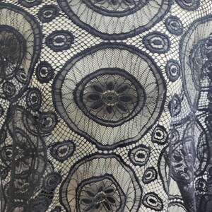 Peut inclure: Un tissu en dentelle noire pr&eacute;sentant un motif r&eacute;p&eacute;titif de grands cercles avec des centres floraux complexes, reli&eacute;s par un filet fin. Le design se compose de bordures circulaires brod&eacute;es et de formes de p&eacute;tales d&eacute;taill&eacute;es sur tout le textile.