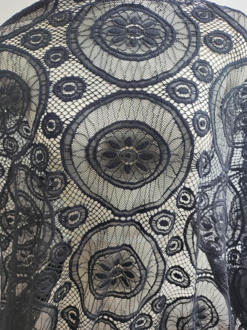Peut inclure: Un tissu en dentelle noire pr&eacute;sentant un motif r&eacute;p&eacute;titif de grands cercles avec des centres floraux complexes, reli&eacute;s par un filet fin. Le design se compose de bordures circulaires brod&eacute;es et de formes de p&eacute;tales d&eacute;taill&eacute;es sur tout le textile.
