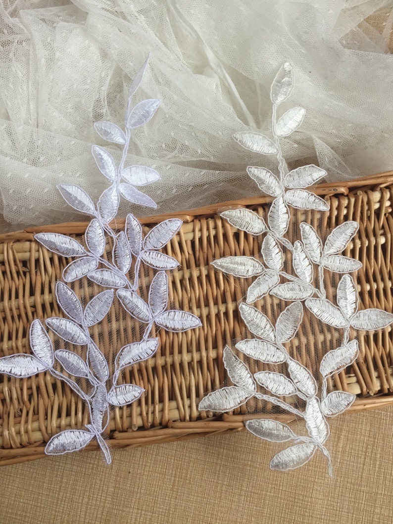 May include: Two white lace appliques in the shape of leafy branches. The appliques are on a woven wicker surface.