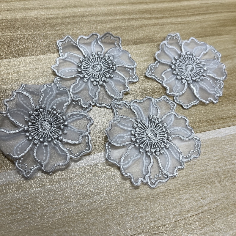 May include: Four individual flower-shaped appliques featuring sheer white organza petals with intricate silver metallic embroidery outlining the edges and a dense, textured silver thread center. Each piece displays a delicate, layered floral design.