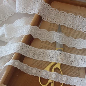 May include: Four white cotton lace trims with scalloped edges and different floral patterns. The lace trims are arranged on a wooden surface.