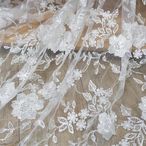 Puede incluir: Primer plano de tela de encaje floral blanco. El delicado encaje presenta flores y hojas bordadas con detalles de lentejuelas. La tela est&aacute; drapeada sobre una superficie de madera, mostrando los intrincados detalles y la textura.