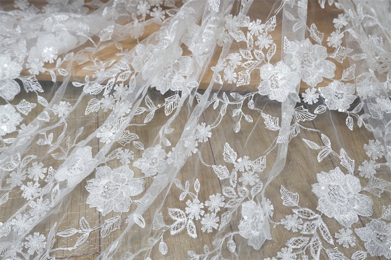 Puede incluir: Primer plano de tela de encaje floral blanco. El delicado encaje presenta flores y hojas bordadas con detalles de lentejuelas. La tela est&aacute; drapeada sobre una superficie de madera, mostrando los intrincados detalles y la textura.