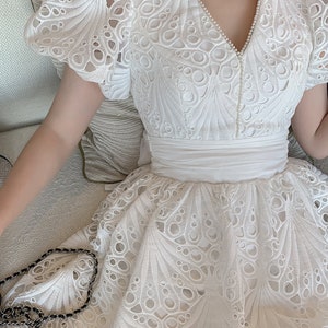Puede incluir: Vestido blanco de encaje con escote en V y mangas cortas abullonadas. El vestido tiene un delicado patr&oacute;n floral y un corpi&ntilde;o ajustado. La falda es amplia y fluida.