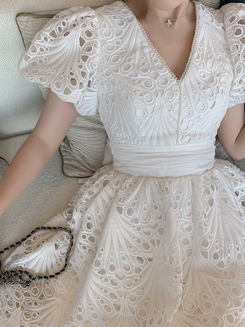 Puede incluir: Vestido blanco de encaje con escote en V y mangas cortas abullonadas. El vestido tiene un delicado patr&oacute;n floral y un corpi&ntilde;o ajustado. La falda es amplia y fluida.
