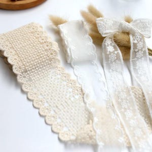 Op de afbeelding: Een collectie kanten linten, waaronder een brede beige kanten rand met schulprand, een smalle witte kanten rand met schulprand en een wit doorschijnend kanten lint met bloemmotief dat is gestrikt tot een strik met twee lange uiteinden.