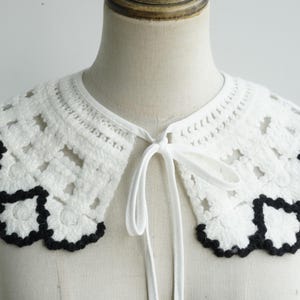 May include: A white crochet collar with a black trim and a tie closure. The collar features a floral pattern and is displayed on a mannequin. The collar is a fashion accessory.