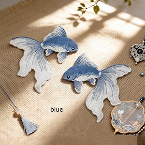 May include: Two embroidered fish patches featuring blue and white thread detailing. The fish have flowing, translucent-looking fins and are positioned on a textured surface. The word blue is printed near the fish. A tassel and a decorative fan are visible.