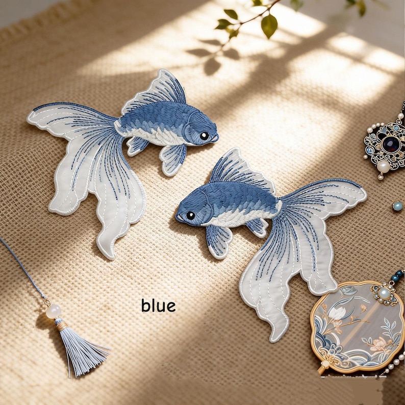 May include: Two embroidered fish patches featuring blue and white thread detailing. The fish have flowing, translucent-looking fins and are positioned on a textured surface. The word blue is printed near the fish. A tassel and a decorative fan are visible.