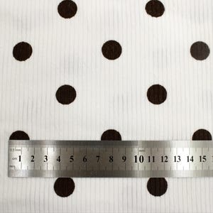 May include: A white corduroy fabric featuring a repeating pattern of dark brown polka dots. A stainless steel ruler is positioned across the bottom, showing the fabric texture and the scale of the dots in centimeters, with markings up to 15 cm.