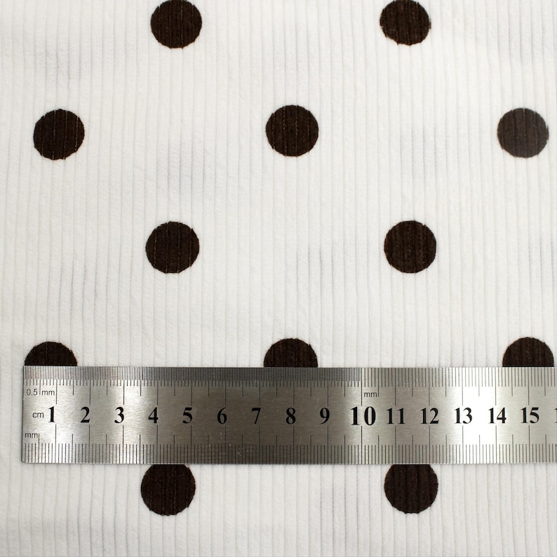 May include: A white corduroy fabric featuring a repeating pattern of dark brown polka dots. A stainless steel ruler is positioned across the bottom, showing the fabric texture and the scale of the dots in centimeters, with markings up to 15 cm.