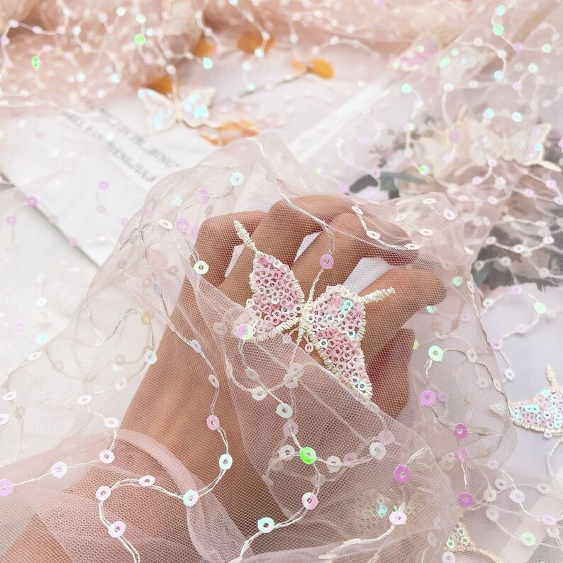 May include: Pink iridescent sequins are sewn onto a sheer pink mesh fabric in the shape of butterflies. The fabric is draped over a hand.