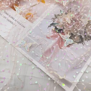 May include: Pink sheer fabric with iridescent sequins and embroidered butterflies. The fabric has a delicate, flowing texture and is perfect for adding a touch of sparkle to any project.