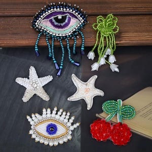 Peut inclure: Une collection de six broches perlées comprenant deux motifs d'œil, l'un aux tons violets et bleus et l'autre blanc et doré, deux étoiles de mer blanches, un bouquet floral en perles vertes et deux cerises rouges à feuilles vertes.