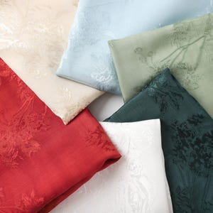May include: A collection of six folded fabric swatches featuring a tonal floral jacquard pattern. The colors include red, white, cream, light blue, sage green, and dark teal, each displaying a woven rose design across the textile surface.