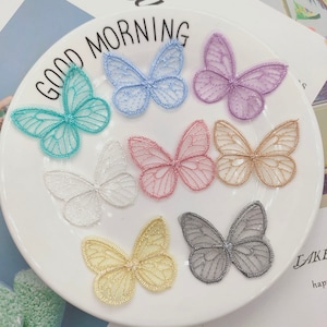 May include: Assortment of eight delicate butterfly-shaped lace appliques in various colors, including light blue, turquoise, purple, pink, gold, white, and gray. The appliques are arranged on a white plate with the words "Good Morning" printed on the edge.