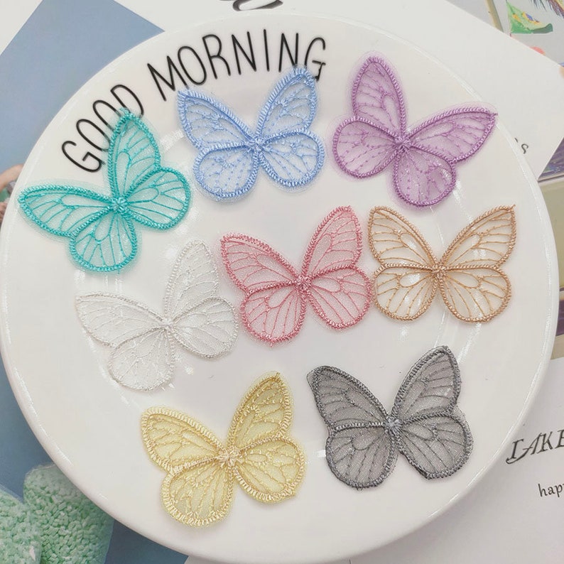 May include: Assortment of eight delicate butterfly-shaped lace appliques in various colors, including light blue, turquoise, purple, pink, gold, white, and gray. The appliques are arranged on a white plate with the words "Good Morning" printed on the edge.