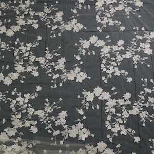 May include: White floral lace fabric featuring a delicate pattern of small flowers and leaves. The lace is sheer and has a soft, flowing texture.