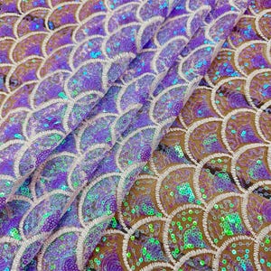 Może przedstawiać: Zbliżenie tkaniny z wzorem łuski syreny. Tkanina ma powtarzający się wzór nakładających się, półkolistych łusek w odcieniach fioletu, turkusu i złota, ozdobionych cekinami i białymi przeszyciami.