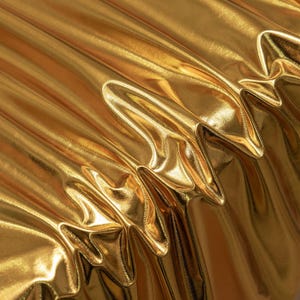 May include: A metallic gold fabric with a high-gloss, reflective finish. The material is draped in deep, vertical folds that create sharp, angular ridges and soft, rounded valleys, highlighting the smooth, liquid-like texture of the surface.