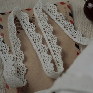 Puede incluir: Borde de encaje blanco de crochet con un delicado borde festoneado. El encaje se muestra doblado en un sobre marrón con marcas rojas y azules.