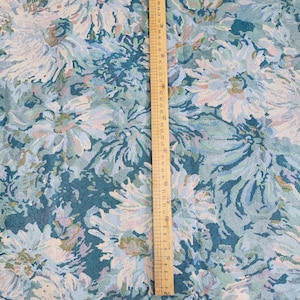 May include: A teal fabric featuring a dense floral pattern in shades of light blue, white, and pale lavender. A wooden ruler is placed vertically on the fabric, showing a measurement scale that extends up to approximately 53 centimeters in length.