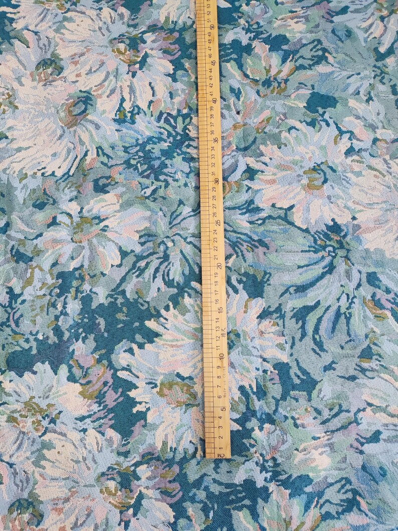 May include: A teal fabric featuring a dense floral pattern in shades of light blue, white, and pale lavender. A wooden ruler is placed vertically on the fabric, showing a measurement scale that extends up to approximately 53 centimeters in length.