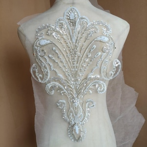May include: A large, symmetrical applique featuring intricate white embroidery, silver metallic thread, and clear sequins arranged in a decorative floral and paisley-inspired pattern on a sheer, light-colored mesh fabric base.