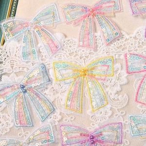 Peut inclure: Une collection d'appliques en forme de nœuds décoratifs en tissu transparent avec des paillettes irisées et des broderies. Chaque nœud est orné de petites perles au centre, présentant une palette de couleurs pastel rose, jaune et bleu.