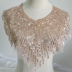 Könnte beinhalten: Ein goldener Paillettenkragen mit rundem Ausschnitt. Der Kragen besteht aus einem durchsichtigen, hellfarbenen Stoff und ist dicht mit kleinen, runden Pailletten bedeckt. Die Pailletten sind in einem Muster angeordnet, das vom Ausschnitt nach außen strahlt.