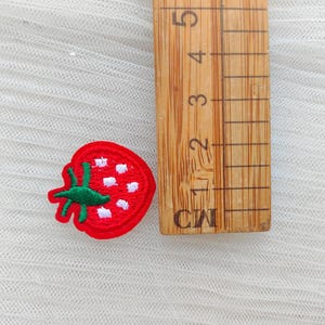 May include: A red embroidered strawberry patch with green leaves and white seeds. A wooden ruler with centimeter markings is next to the patch. The patch is on a white, textured surface.