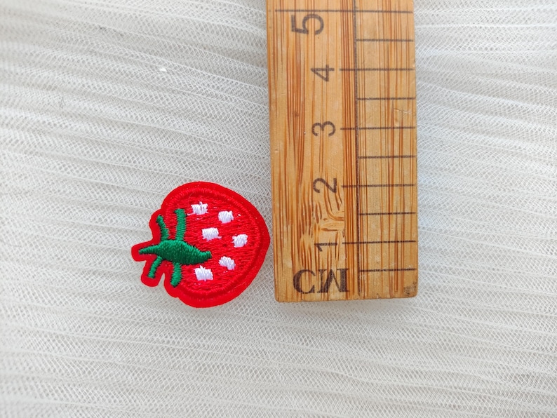 May include: A red embroidered strawberry patch with green leaves and white seeds. A wooden ruler with centimeter markings is next to the patch. The patch is on a white, textured surface.