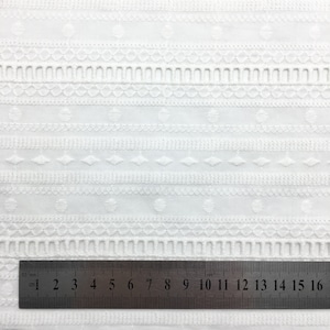 May include: White cotton fabric with an embroidered pattern of small circles, diamonds, and lines. A ruler is placed at the bottom of the image, showing measurements in inches.