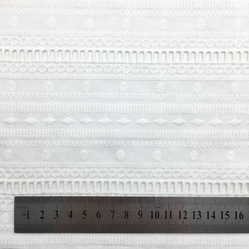 May include: White cotton fabric with an embroidered pattern of small circles, diamonds, and lines. A ruler is placed at the bottom of the image, showing measurements in inches.