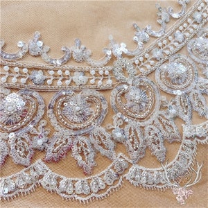 Pu&ograve; includere: Primo piano di un bordino in pizzo bianco perlato e paillettes. Il bordino presenta un delicato disegno floreale con perline madreperlacee e paillettes argentate.