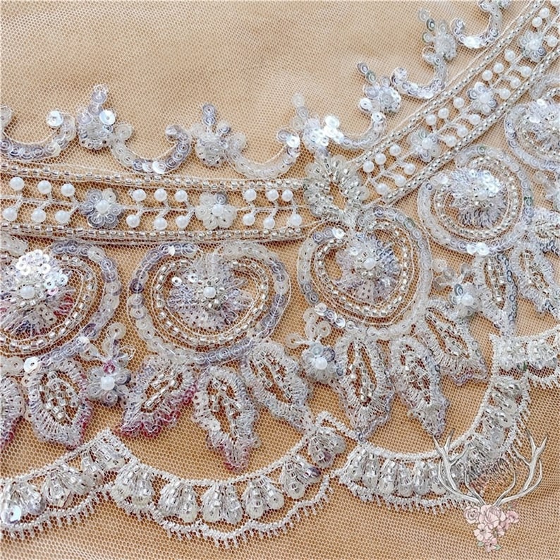Pu&ograve; includere: Primo piano di un bordino in pizzo bianco perlato e paillettes. Il bordino presenta un delicato disegno floreale con perline madreperlacee e paillettes argentate.