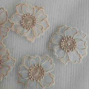May include: Three floral appliques featuring sheer white organza petals with scalloped edges and intricate beige embroidery detailing the flower centers and petal outlines, arranged on a fine white mesh fabric background.