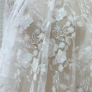 Puede incluir: Tela de encaje floral blanco con flores y hojas bordadas. El delicado dise&ntilde;o presenta costuras detalladas y un fondo transparente, creando un aspecto texturizado y elegante. Ideal para vestidos de novia, vestidos o proyectos decorativos.