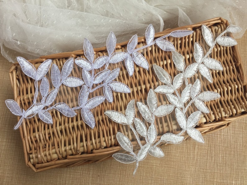 May include: Two silver leaf lace appliques with a delicate floral design. The appliques are on a woven wicker surface.
