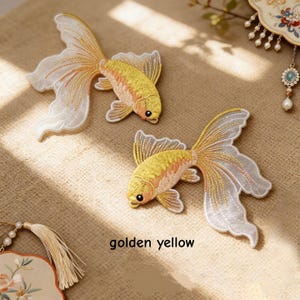 May include: Two embroidered goldfish patches featuring golden yellow bodies with intricate scale detailing and long, flowing white translucent fins. The patches are positioned diagonally, and the text golden yellow is printed below them.