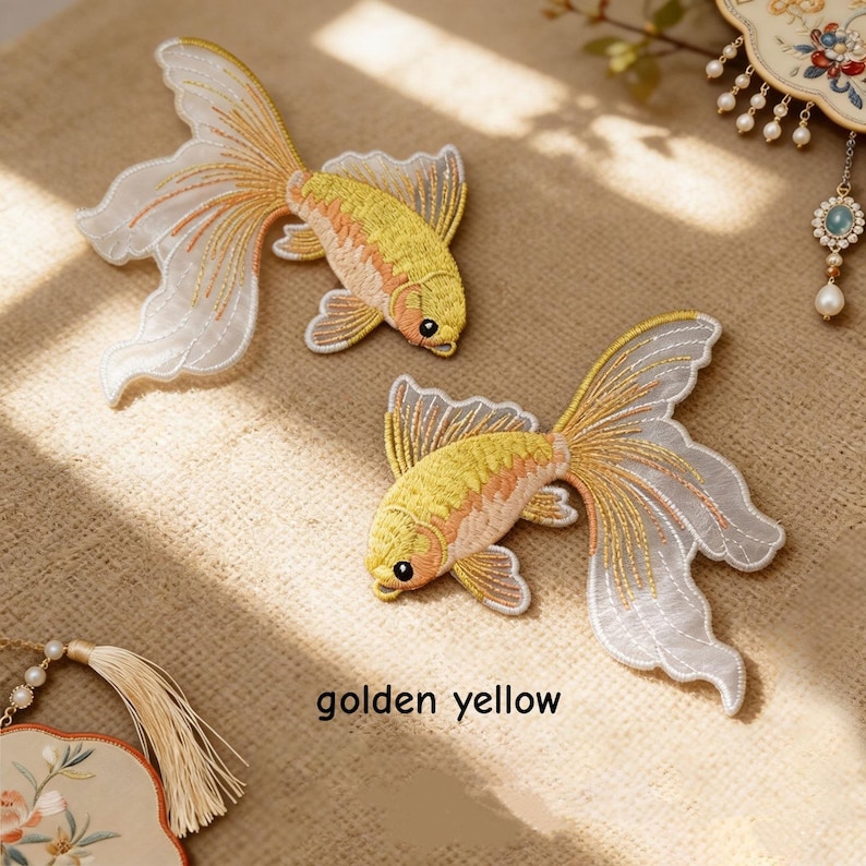 May include: Two embroidered goldfish patches featuring golden yellow bodies with intricate scale detailing and long, flowing white translucent fins. The patches are positioned diagonally, and the text golden yellow is printed below them.
